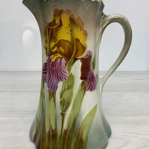 Puede incluir: Una jarra de cerámica con una flor de iris pintada con pétalos amarillos y morados sobre un fondo gris claro. La jarra tiene un borde festoneado y un asa curva, ambos acentuados con un fino borde dorado a lo largo de los bordes y el pico.