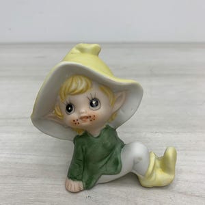 May include: A ceramic elf figurine wearing a large, wide-brimmed yellow hat, a green long-sleeved shirt, and yellow boots. The elf has blonde hair, pointed ears, and freckles on the cheeks, depicted in a seated position with legs extended.
