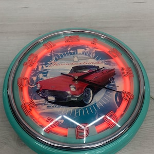 May include: A round wall clock with a teal frame and a chrome bezel. The clock face features a red Thunderbird car illustration with the text "Thunderbird" and a bright red neon light ring. It has black hour, minute, and second hands.