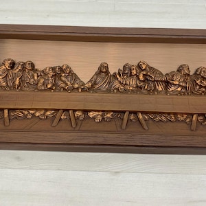 May include: A rectangular wall plaque featuring a copper-toned relief sculpture of The Last Supper. The scene depicts thirteen figures seated at a long table, framed within a dark brown wooden border. The metallic artwork has a polished finish.