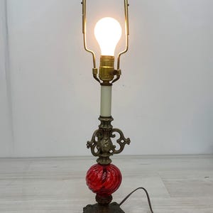May include: A table lamp featuring a brass base with ornate scrollwork, a central red glass sphere with a twisted ribbed texture, and a brass harp. The lamp includes a white light bulb and a brown electrical cord extending from the base.