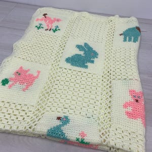 May include: A cream-colored crochet blanket featuring a grid of square panels. Each panel displays a cross-stitched animal motif in pastel pink or teal, including a rabbit, a cat, a bird, and a bear, separated by open-work crochet sections.