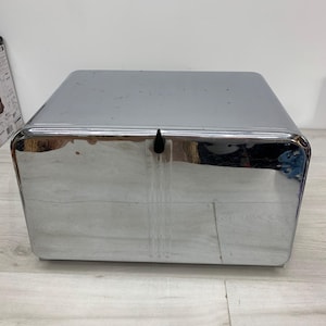 May include: A rectangular bread box with a polished chrome finish and a reflective surface. The container features a hinged front door with a small black handle centered at the top edge. The metal surface shows minor signs of wear and surface scratches.