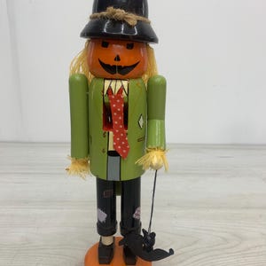 May include: A wooden nutcracker figure designed as a scarecrow with a pumpkin head, wearing a black top hat, a green jacket, and black pants. The figure features a red polka-dot tie, yellow raffia hair, and holds a small black bat on a string.