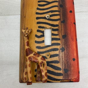 May include: A rectangular wooden light switch plate featuring a hand-carved, three-dimensional giraffe figure on the left side. The background is painted with a yellow section, a central black zebra-striped pattern, and a red-to-brown gradient edge.