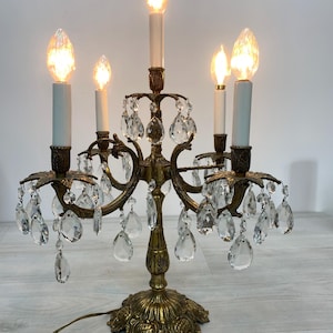 Puede incluir: Una lámpara candelabro de cinco luces con base y estructura de metal en tono latón. La lámpara cuenta con cinco fundas blancas estilo vela, cada una coronada con una bombilla encendida. Numerosos prismas de cristal transparente en forma de gota.