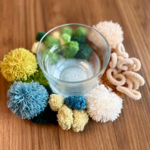 May include: A clear glass filled with water sits on a colorful, handmade coaster. The coaster is adorned with various pom-poms in shades of yellow, blue, green, and cream, creating a textured and decorative base for the glass.