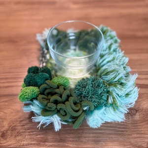 Op de afbeelding: Een handgemaakte kandelaar met een heldere glazen beker in het midden. De houder is omgeven door groene en lichtblauwe wol, met verschillende texturen en groentinten, wat een uniek decoratief stuk creëert.