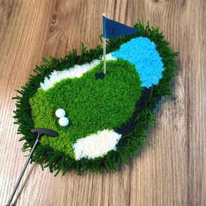May include: A handcrafted golf-themed rug featuring a green putting green, a blue water hazard, and a white sand trap. Includes two golf balls, a miniature flag, and a putter. The rug is made of textured yarn.