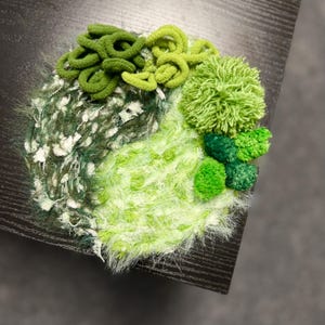 May include: A textured, abstract textile art piece in shades of green and white. The design features various textures, including looped yarn, fluffy fibers, and small, round, pom-pom-like elements, creating a layered, three-dimensional effect. The piece is set against a dark background.