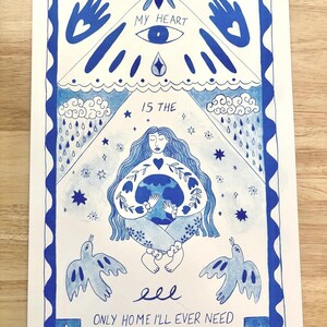 Op de afbeelding: Blauw-witte illustratie met de tekst "My Heart Is The Only Home I'll Ever Need". Het kunstwerk toont een vrouw die de aarde omarmt, omringd door hemelse en natuurlijke elementen, waaronder vogels, sterren en regen.