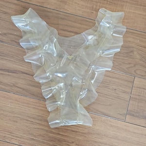 May include: A translucent, ruffled collar-like accessory, possibly for a costume or garment. The material appears to be a thin, flexible plastic or similar synthetic. The item is laid on a wooden surface.