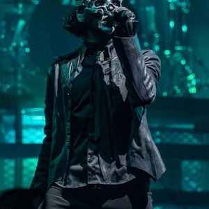 May include: A performer wearing a silver skull mask with a black leather jacket and black shirt. The performer is on stage, with a dark background and stage lighting. The mask has intricate details and is the focal point of the image.