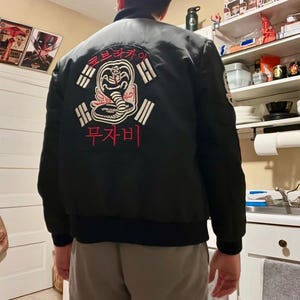 Op de afbeelding: Zwarte bomberjack met een Cobra Kai-logo geborduurd op de rug. Het logo toont een cobra met Koreaanse tekst. De jas heeft een geribbelde kraag, manchetten en zoom. De jas wordt door een persoon gedragen.