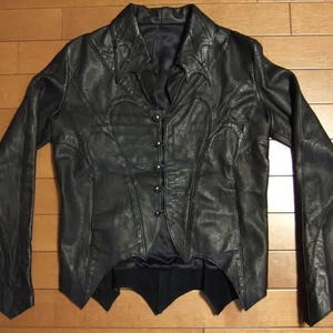 May include: Black leather jacket with a unique design. The jacket features a button-up front, a notched collar, and decorative stitching. The sleeves are long and flared, and the jacket has a tailored fit. The jacket is displayed on a wooden surface.
