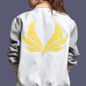 Op de afbeelding: Witte en grijze varsity jas met een gele geborduurde vleugel op de rug. De jas heeft gele en witte gestreepte randen op de kraag, manchetten en taille. De mouwen zijn grijs en de body is wit.