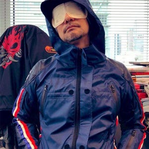 Op de afbeelding: Een marineblauwe jas met capuchon, zwarte details en een ritssluiting. De mouwen hebben rood-wit-blauwe strepen. De capuchon heeft witte tekst. De persoon draagt een wit oogmasker.