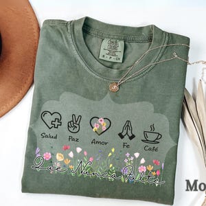 Puede incluir: Camiseta Comfort Colors verde musgo con un dise&ntilde;o floral y las palabras "Que Nunca Falte". El dise&ntilde;o incluye s&iacute;mbolos de salud, paz, amor, fe y caf&eacute;. Tambi&eacute;n se ven un collar dorado y un sombrero marr&oacute;n.