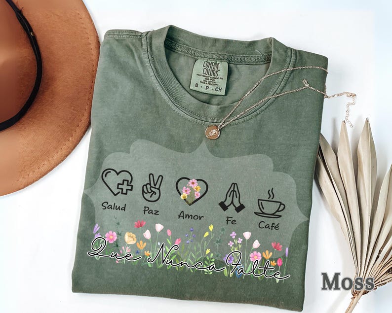 Puede incluir: Camiseta Comfort Colors verde musgo con un dise&ntilde;o floral y las palabras "Que Nunca Falte". El dise&ntilde;o incluye s&iacute;mbolos de salud, paz, amor, fe y caf&eacute;. Tambi&eacute;n se ven un collar dorado y un sombrero marr&oacute;n.