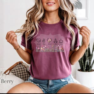 Puede incluir: Camiseta color bayas con un dise&ntilde;o floral en la parte inferior y las palabras "Salud, Paz, Amor, Fe, Caf&eacute;" encima de las flores. La camiseta tiene mangas cortas y la lleva una persona.