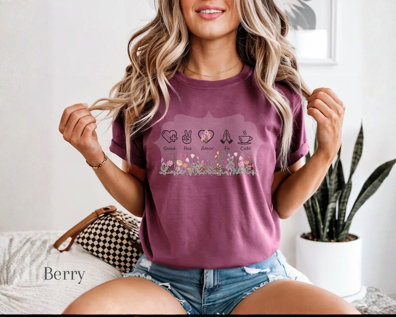 Puede incluir: Camiseta color bayas con un dise&ntilde;o floral en la parte inferior y las palabras "Salud, Paz, Amor, Fe, Caf&eacute;" encima de las flores. La camiseta tiene mangas cortas y la lleva una persona.