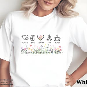 Puede incluir: Camiseta blanca con un dise&ntilde;o gr&aacute;fico que presenta s&iacute;mbolos de salud, paz, amor, fe y caf&eacute;. El dise&ntilde;o incluye elementos florales y el texto "Que Nunca Falte". La camiseta es de cuello redondo.