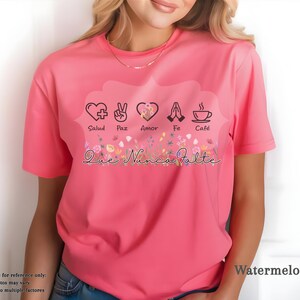 Puede incluir: Camiseta de color sand&iacute;a con un dise&ntilde;o gr&aacute;fico que incluye un coraz&oacute;n, un signo de la paz, un coraz&oacute;n, manos rezando y una taza de caf&eacute;. El texto "Salud, Paz, Amor, Fe, Caf&eacute;" est&aacute; encima de los s&iacute;mbolos y "Que Nunca Falte" debajo.