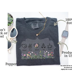 Puede incluir: Camiseta gris oscuro con dise&ntilde;o floral y las palabras "Salud, Paz, Amor, Fe, Caf&eacute;". Hecha de 100% algod&oacute;n estadounidense y producida en EE. UU. Tambi&eacute;n se ven un collar dorado y gafas de sol.