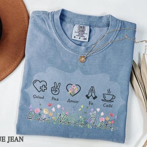 Puede incluir: Camiseta azul vaquero Comfort Colors con un dise&ntilde;o floral y las palabras "Salud, Paz, Amor, Fe, Caf&eacute;" y "Que Nunca Falte". Un collar dorado y un sombrero marr&oacute;n est&aacute;n junto a la camiseta.