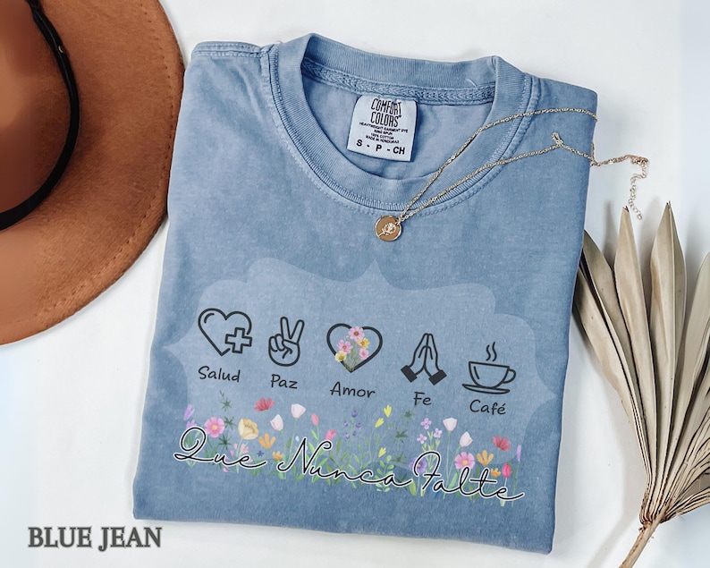 Puede incluir: Camiseta azul vaquero Comfort Colors con un dise&ntilde;o floral y las palabras "Salud, Paz, Amor, Fe, Caf&eacute;" y "Que Nunca Falte". Un collar dorado y un sombrero marr&oacute;n est&aacute;n junto a la camiseta.