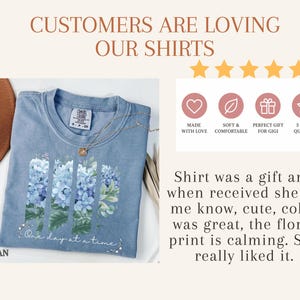 May include: A blue t-shirt featuring a graphic of blue hydrangea flowers arranged in three vertical panels with the text "One day at a time" below. The image includes text stating "CUSTOMERS ARE LOVING OUR SHIRTS" and a five-star review summary.