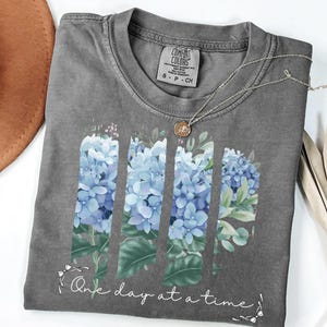 May include: A charcoal gray crew neck t-shirt featuring a graphic of blue hydrangea flowers arranged in three vertical panels. Below the flowers, the text "One day at a time" is printed in a white script font, accented by small white bows.