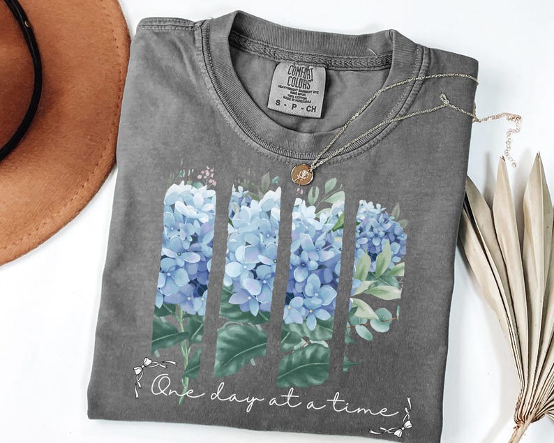May include: A charcoal gray crew neck t-shirt featuring a graphic of blue hydrangea flowers arranged in three vertical panels. Below the flowers, the text "One day at a time" is printed in a white script font, accented by small white bows.
