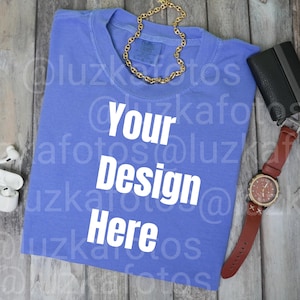 Puede incluir: Una camiseta azul bígaro con el texto "Your Design Here" en blanco. Una cadena dorada, una cartera negra y un reloj de pulsera con una correa de cuero marrón están dispuestos sobre una superficie de madera.