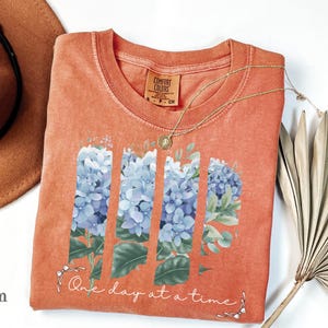 May include: A terracotta-colored t-shirt featuring a graphic of blue hydrangea flowers arranged in four vertical panels. Below the flowers, the text reads One day at a time in white cursive script, accented by small white bows at each end.