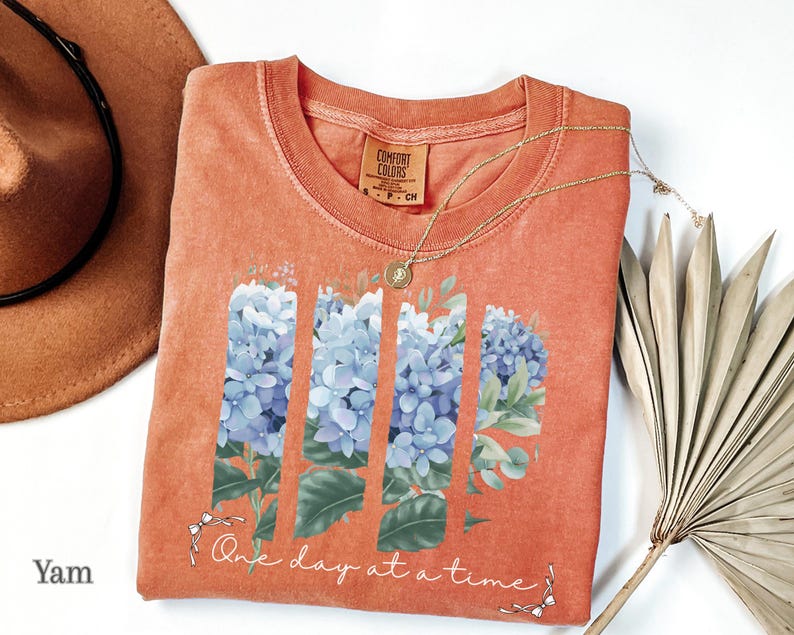 May include: A terracotta-colored t-shirt featuring a graphic of blue hydrangea flowers arranged in four vertical panels. Below the flowers, the text reads One day at a time in white cursive script, accented by small white bows at each end.