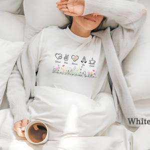Puede incluir: Camiseta blanca de manga larga con dise&ntilde;o floral y las palabras "Salud, Paz, Amor, Fe, Caf&eacute;" y "Dios Nunca Falta". La camiseta tiene cuello redondo y es de material suave.