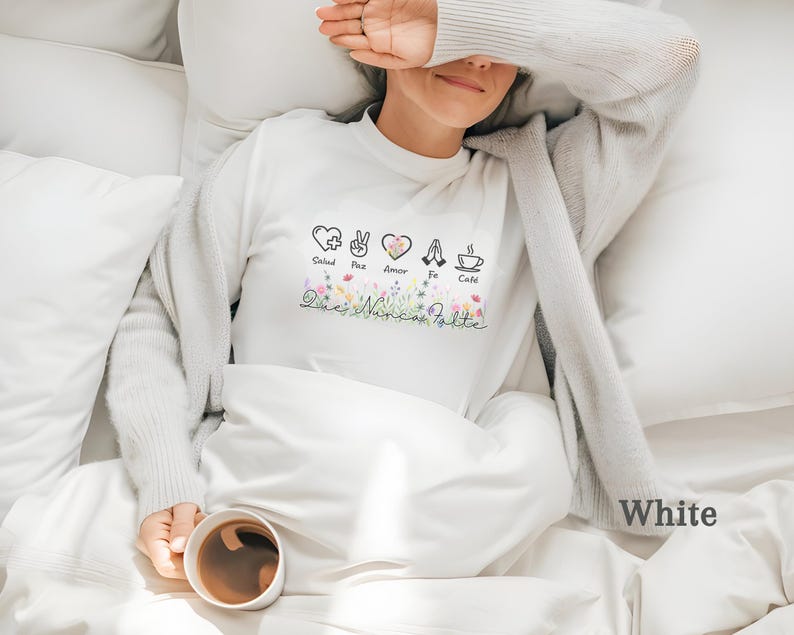 Puede incluir: Camiseta blanca de manga larga con dise&ntilde;o floral y las palabras "Salud, Paz, Amor, Fe, Caf&eacute;" y "Dios Nunca Falta". La camiseta tiene cuello redondo y es de material suave.