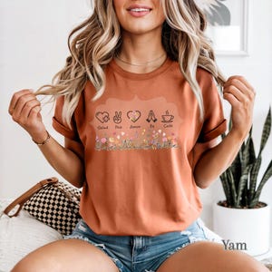 Puede incluir: Camiseta color &oacute;xido con un dise&ntilde;o floral y las palabras "Salud, Paz, Amor, Fe, Caf&eacute;" sobre una escritura cursiva "Vanessa Valiente". La camiseta presenta varios s&iacute;mbolos, incluyendo un coraz&oacute;n, un signo de la paz, manos rezando y una taza de caf&eacute;.
