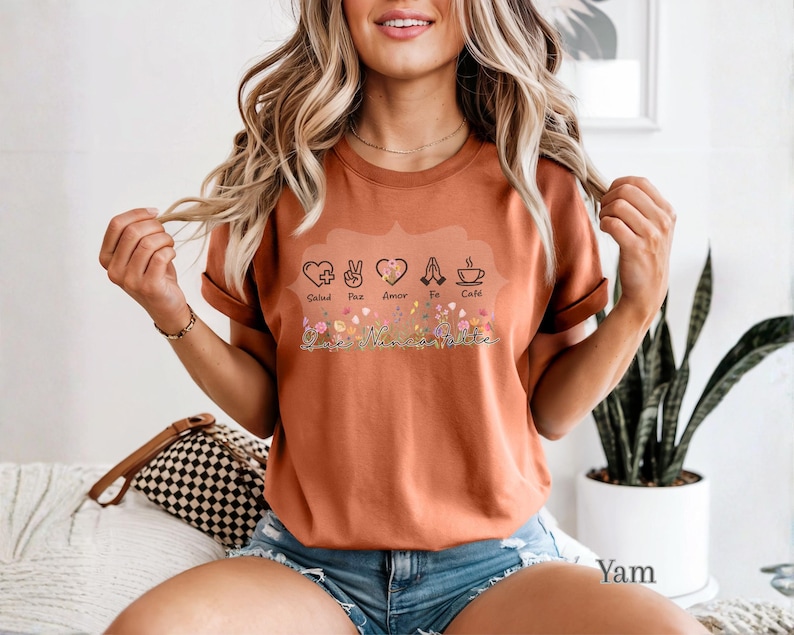 Puede incluir: Camiseta color &oacute;xido con un dise&ntilde;o floral y las palabras "Salud, Paz, Amor, Fe, Caf&eacute;" sobre una escritura cursiva "Vanessa Valiente". La camiseta presenta varios s&iacute;mbolos, incluyendo un coraz&oacute;n, un signo de la paz, manos rezando y una taza de caf&eacute;.