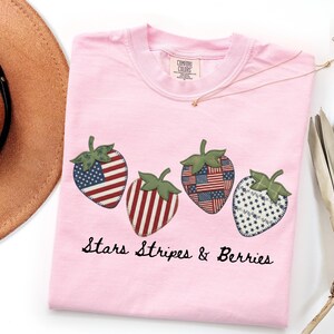 Puede incluir: Una camiseta rosa claro con cuatro diseños de fresas con patrones patrióticos. Las fresas tienen copas verdes y están decoradas con la bandera estadounidense y estrellas. El texto "Stars Stripes & Berries" está impreso debajo.