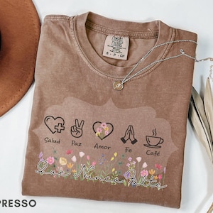 Puede incluir: Camiseta color espresso con un gr&aacute;fico que presenta un coraz&oacute;n, un signo de paz, un coraz&oacute;n con flores, manos rezando y una taza de caf&eacute;. La camiseta tiene las palabras "Salud, Paz, Amor, Fe, Caf&eacute;" y "Que Nunca Falte". Un collar plateado descansa sobre la camiseta.