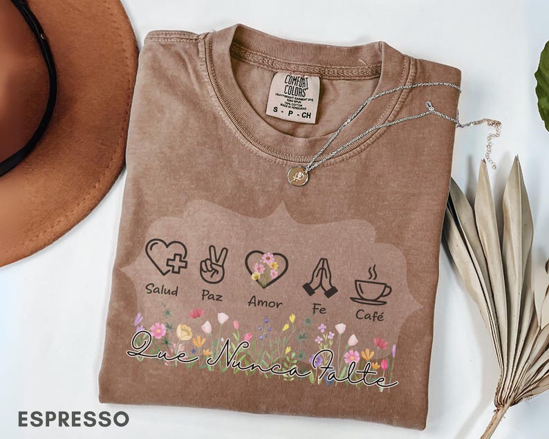 Puede incluir: Camiseta color espresso con un gr&aacute;fico que presenta un coraz&oacute;n, un signo de paz, un coraz&oacute;n con flores, manos rezando y una taza de caf&eacute;. La camiseta tiene las palabras "Salud, Paz, Amor, Fe, Caf&eacute;" y "Que Nunca Falte". Un collar plateado descansa sobre la camiseta.