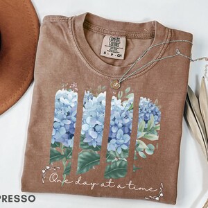 May include: A brown short-sleeve t-shirt featuring a graphic of three vertical panels of blue hydrangea flowers and green leaves. Below the floral design, the text reads One day at a time in white cursive script. The shirt has a crew neckline.