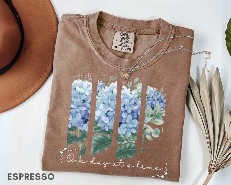 May include: A brown short-sleeve t-shirt featuring a graphic of three vertical panels of blue hydrangea flowers and green leaves. Below the floral design, the text reads One day at a time in white cursive script. The shirt has a crew neckline.