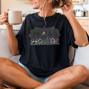 Puede incluir: Camiseta negra con un dise&ntilde;o floral y las palabras "Calm, Peace, Amor, Fe, Cafe". El dise&ntilde;o incluye una fila de flores. La camiseta tiene un corte holgado y mangas cortas.
