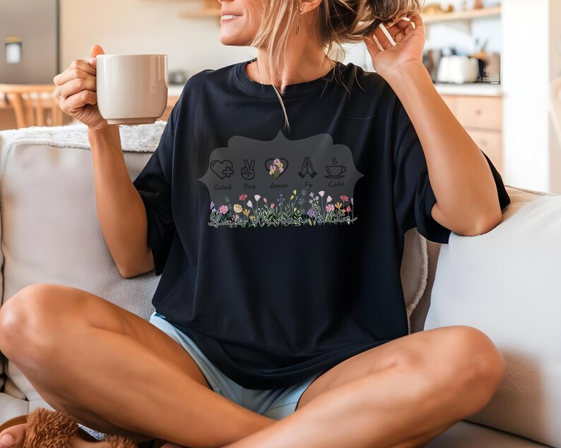 Puede incluir: Camiseta negra con un dise&ntilde;o floral y las palabras "Calm, Peace, Amor, Fe, Cafe". El dise&ntilde;o incluye una fila de flores. La camiseta tiene un corte holgado y mangas cortas.