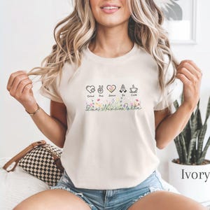 Puede incluir: Camiseta de color marfil con un dise&ntilde;o gr&aacute;fico que incluye las palabras Salud, Paz, Amor, Fe y Caf&eacute;, y detalles florales. El dise&ntilde;o incluye iconos de coraz&oacute;n, signo de la paz y taza de caf&eacute;. La camiseta es de material suave.