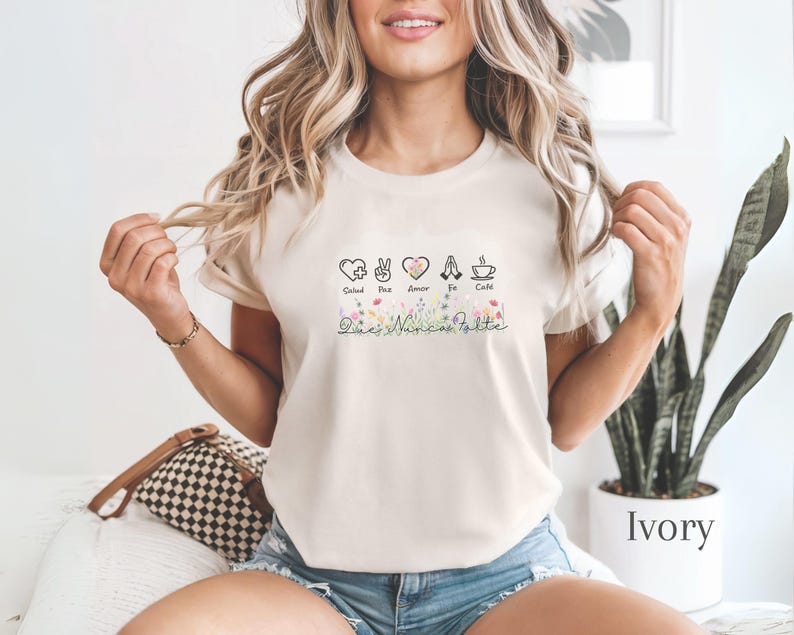 Puede incluir: Camiseta de color marfil con un dise&ntilde;o gr&aacute;fico que incluye las palabras Salud, Paz, Amor, Fe y Caf&eacute;, y detalles florales. El dise&ntilde;o incluye iconos de coraz&oacute;n, signo de la paz y taza de caf&eacute;. La camiseta es de material suave.
