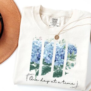 May include: A cream-colored t-shirt featuring a graphic of blue hydrangea flowers arranged in three vertical panels. Below the flowers, the text "One day at a time" is printed in a black script font, accented by small line-drawn bows on each side.